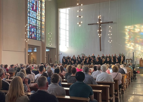 Bild: Publikum sitzend in vollbesetzter Kirche, dahinter singender Chor.