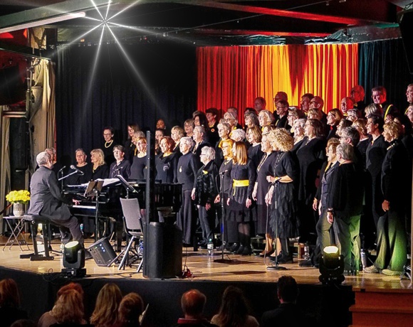 Bild: Auf der B�hne Chorleiter am Klavier spielend und der Chor von Scheinwerfern angestrahlt vor rot, gelb, gr�n und blauen Vorh�ngen singend.