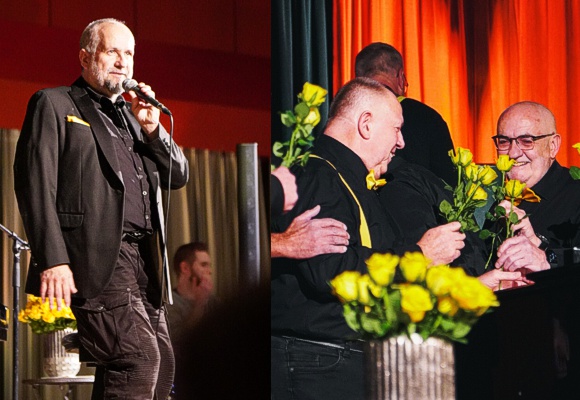 Bild: Links Chorleiter zum Publikum sprechend, rechts Chormitglied Theo, der mit Rosen von den Mitgliedern beschenkt wird.